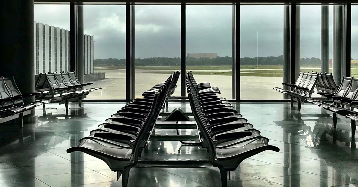 Empty airport waiting hall with rows of black seats facing tall windows that look out on a runway under a cloudy sky