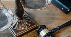 Wooden scales of justice and a black and gold gavel resting on a wooden table beside a dark leather binder