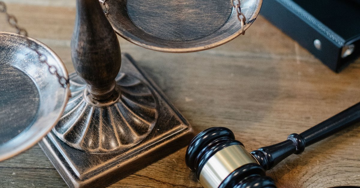 Wooden scales of justice and a black and gold gavel resting on a wooden table beside a dark leather binder