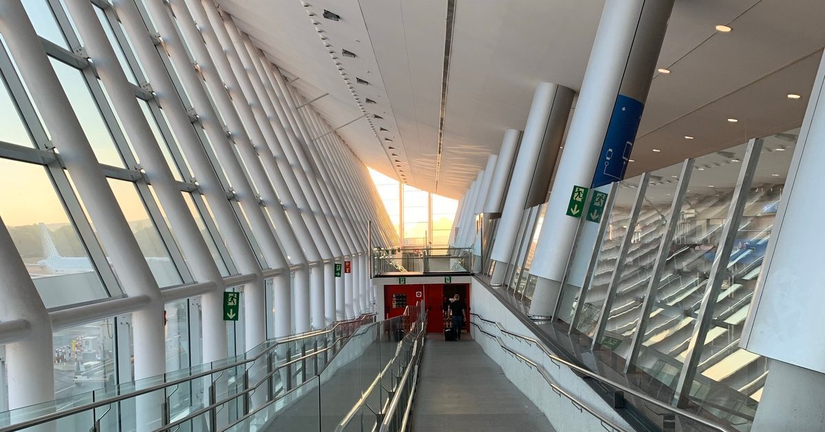 Long glass-walled airport walkway at sunrise, with a lone traveler and a suitcase far in the distance heading toward the terminal